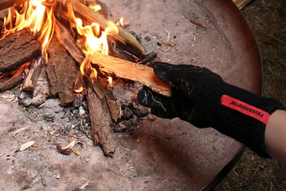 Hand mit Handschuh hält Holzscheit ins Feuer